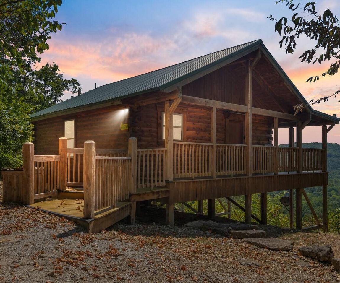 A log cabin with a covered wraparound porch on a wooded ridge at sunset, looking out over the Ozark hills.