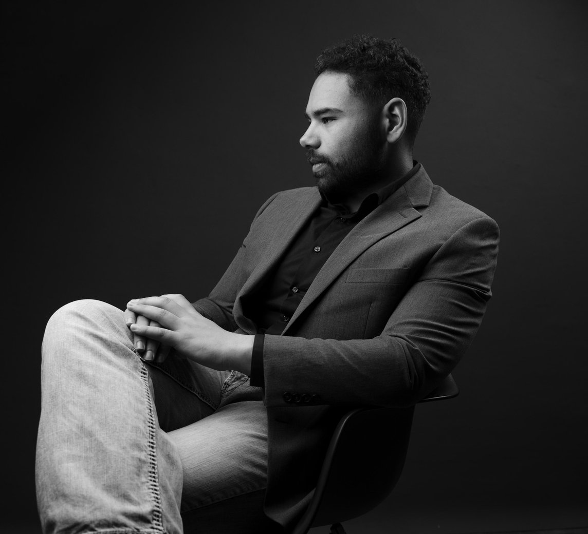 Devin Osborne seated in a chair, looking off to the side, in a grey blazer and jeans.
